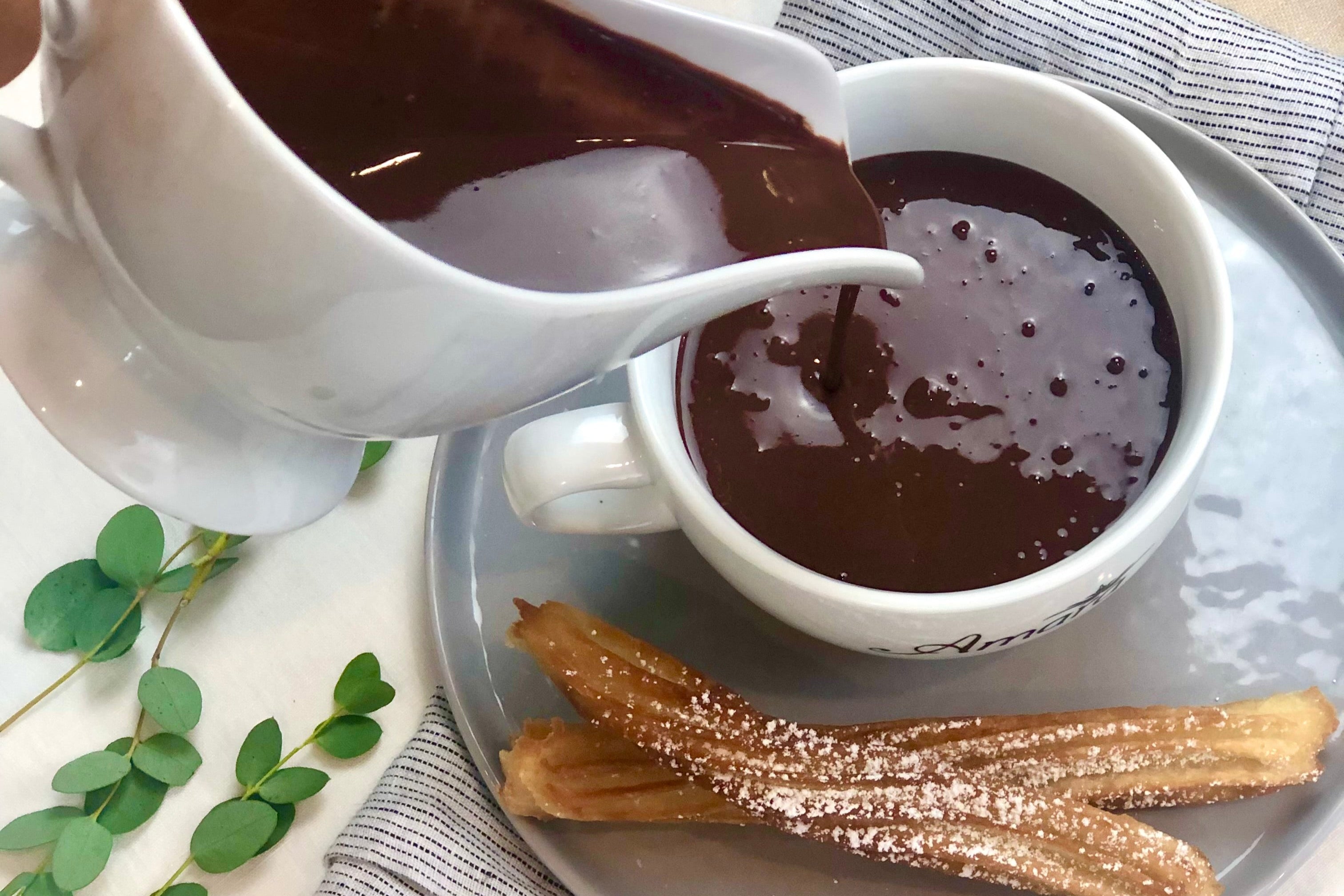 Best Hot Chocolate being poured from a white pitcher into a white mug with churros on a light wooden surface